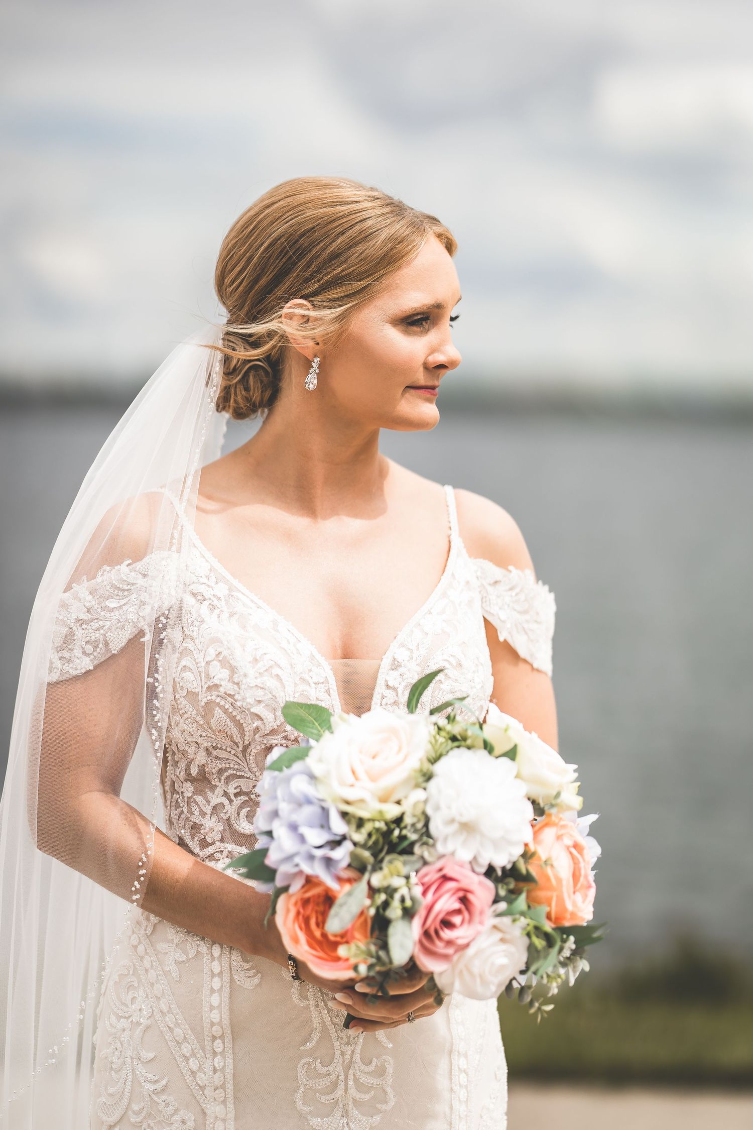 Bride wearing delicate lace gown and veil holds pastel bouquet by waterfront on cloudy day.
