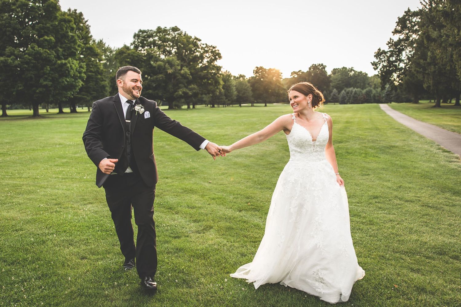 A couple runs playfully across a green lawn during their wedding photoshoot at sunset.