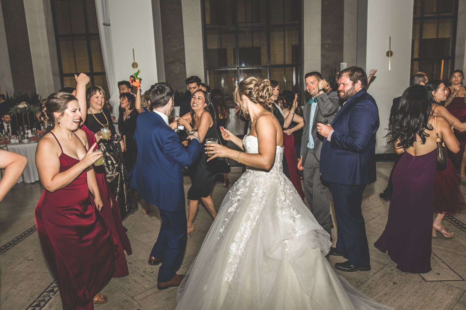 Wedding party and guests dancing in formal attire at an elegant indoor reception venue.