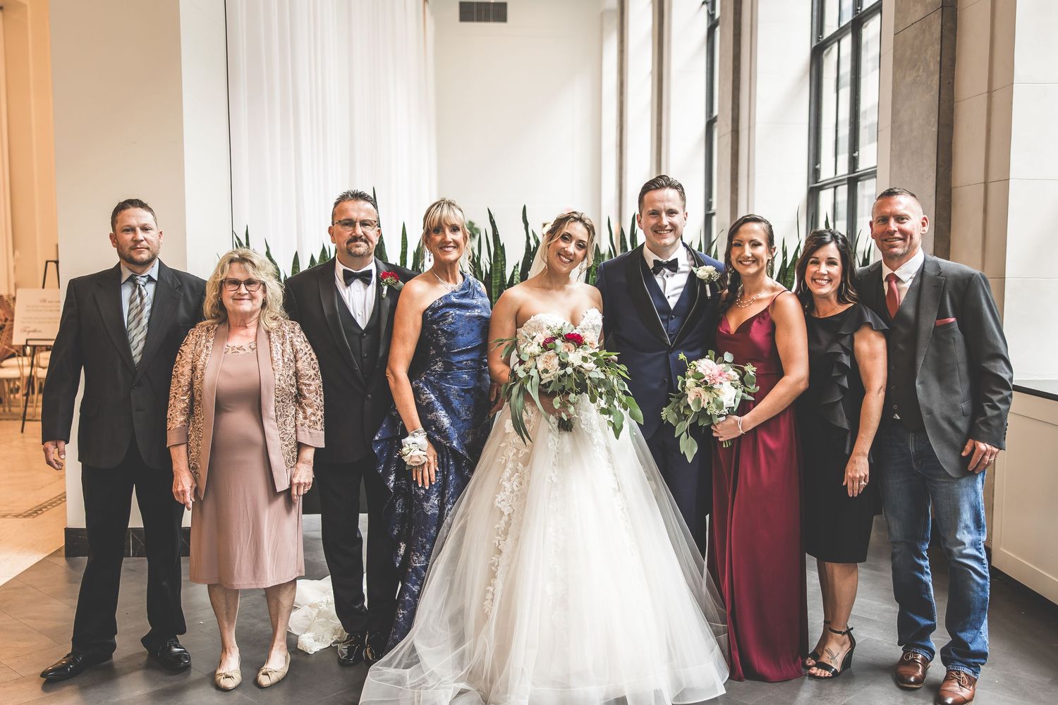Full wedding party poses together in formal attire along bright hallway.