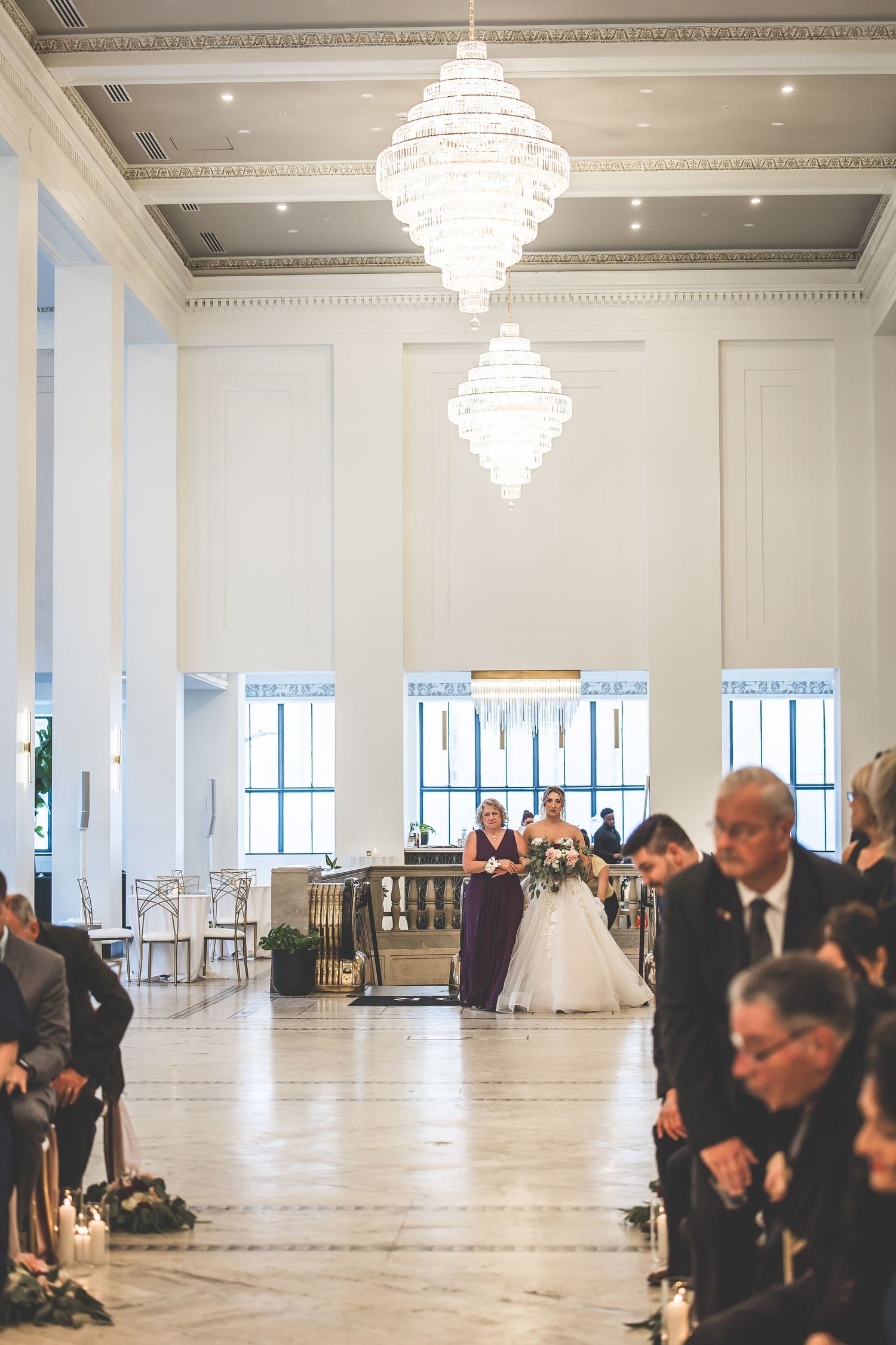 Wedding ceremony in an elegant white ballroom with crystal chandeliers and guests seated in rows.