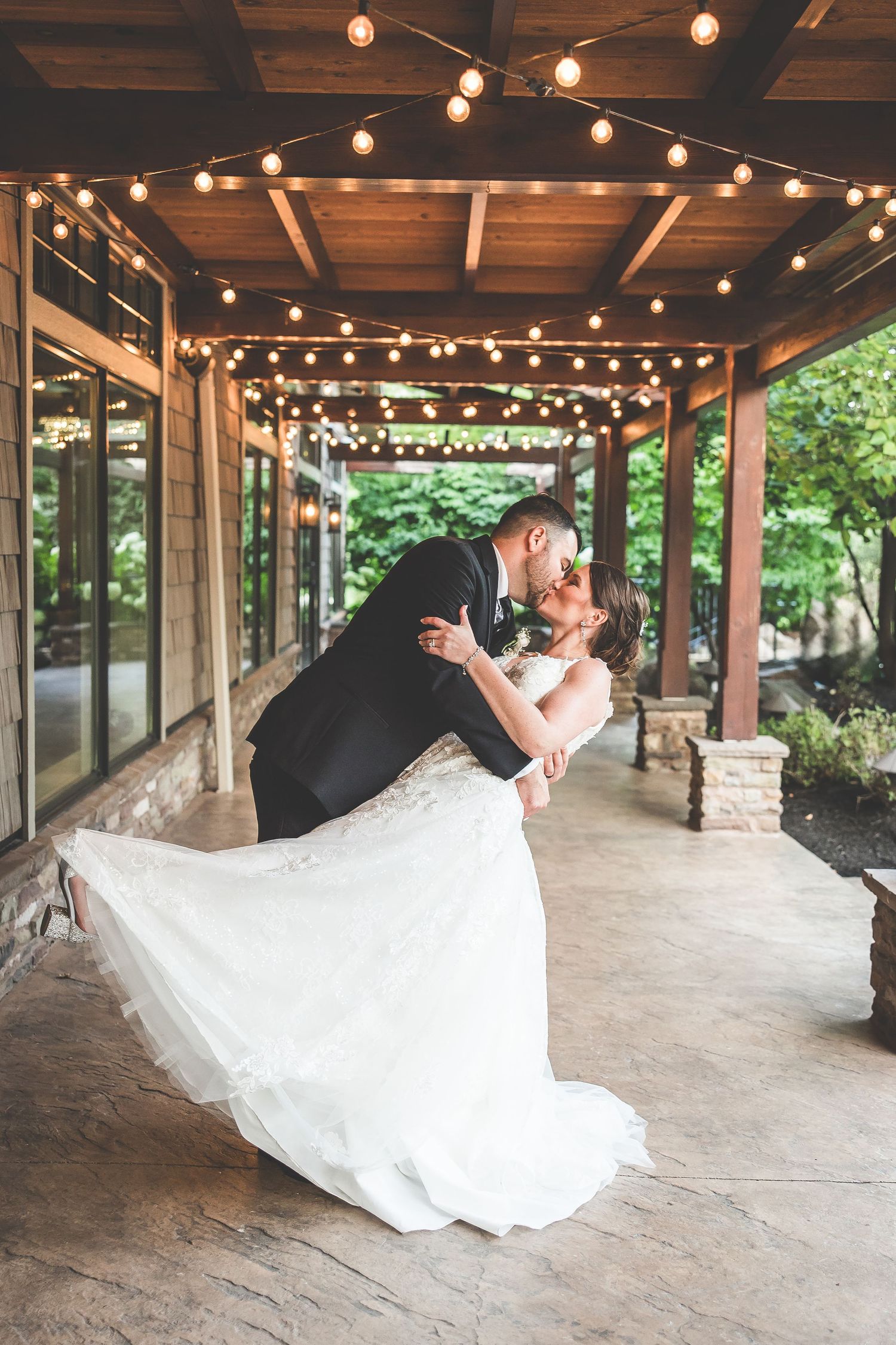 Dramatic wedding dip pose under string lights in rustic outdoor pavilion.