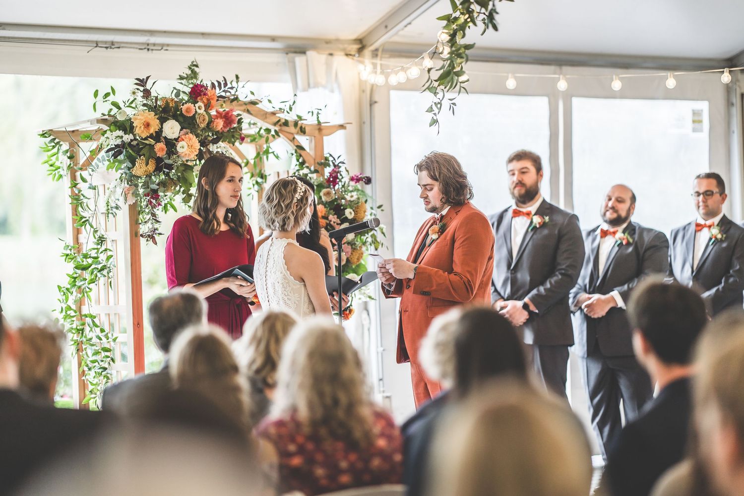 Wedding ceremony in a bright indoor venue with floral arch decorations and guests seated in rows.
