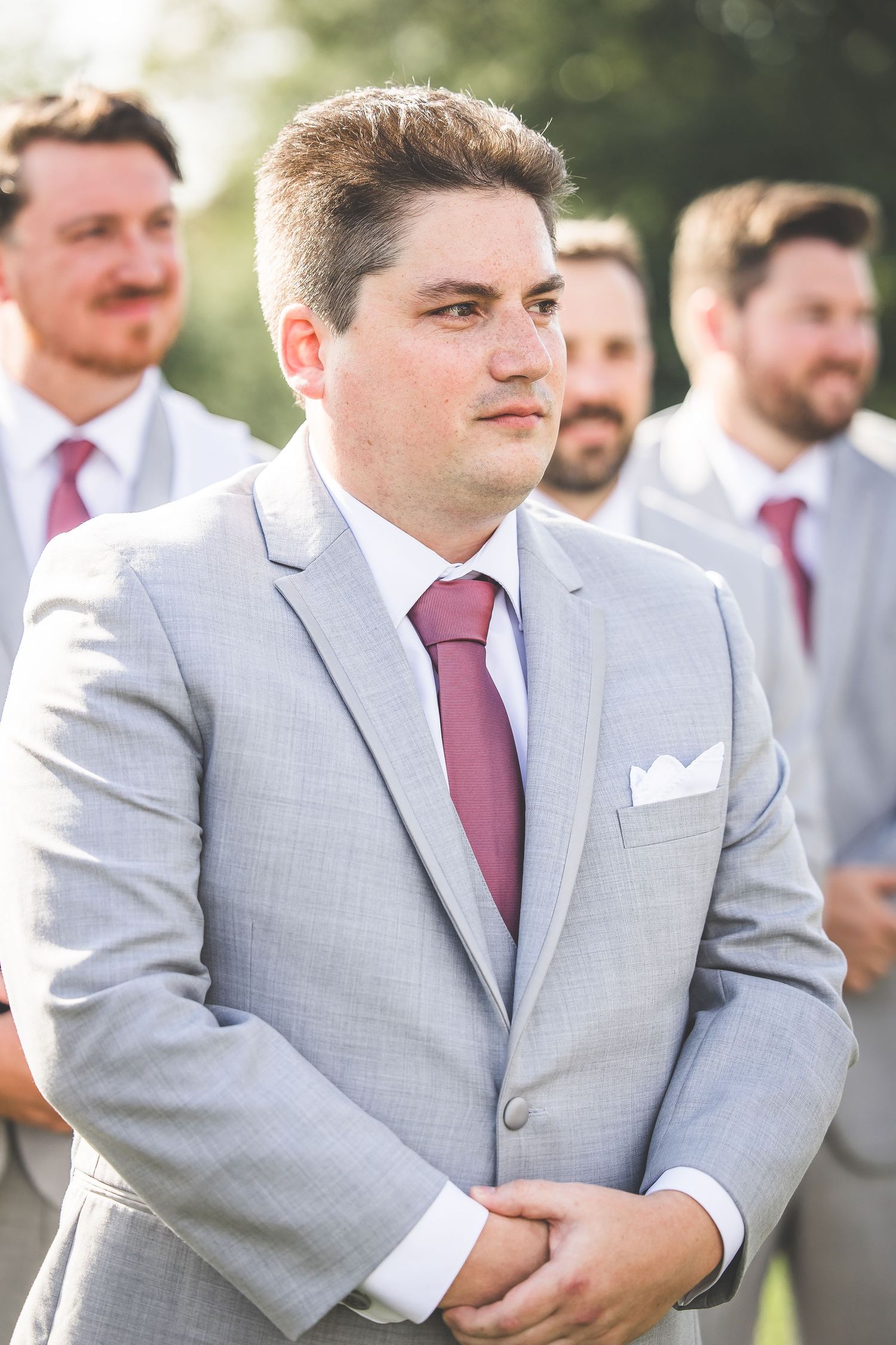 Groomsman in light grey suit with burgundy tie stands during outdoor wedding ceremony.