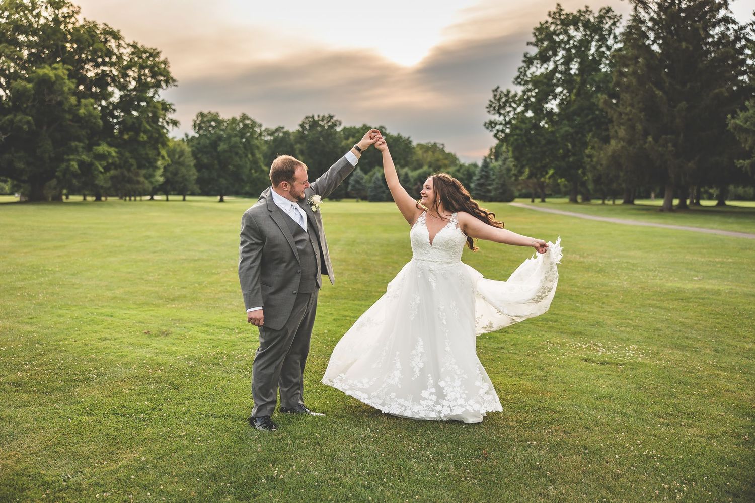 Newlyweds dancing and twirling on expansive green lawn at sunset.