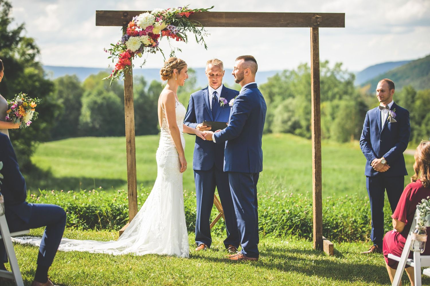 Couple exchanging vows under rustic wooden arch with floral decorations in scenic outdoor setting.