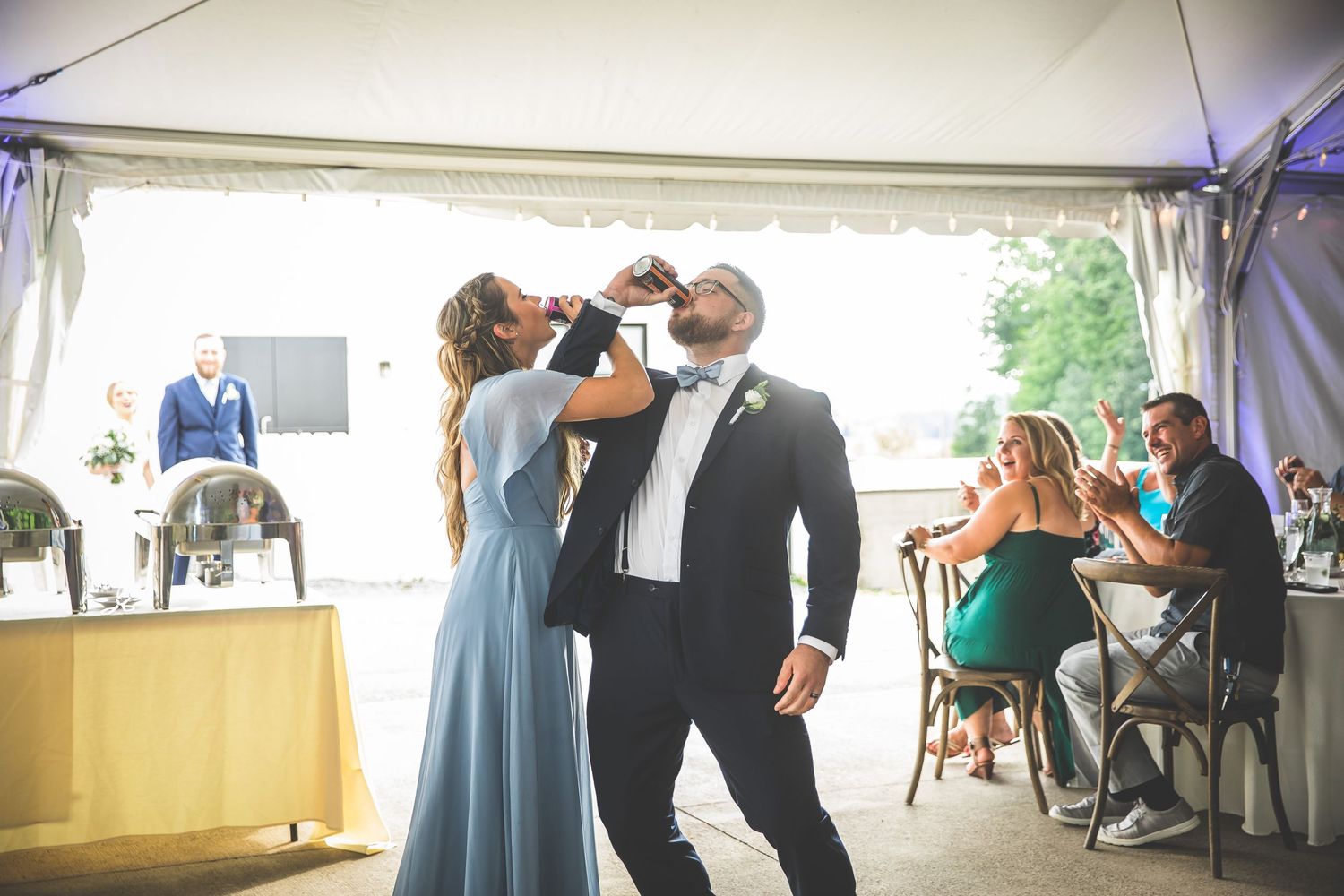 A couple shares their first dance at a wedding reception under a white tent with string lights and greenery.