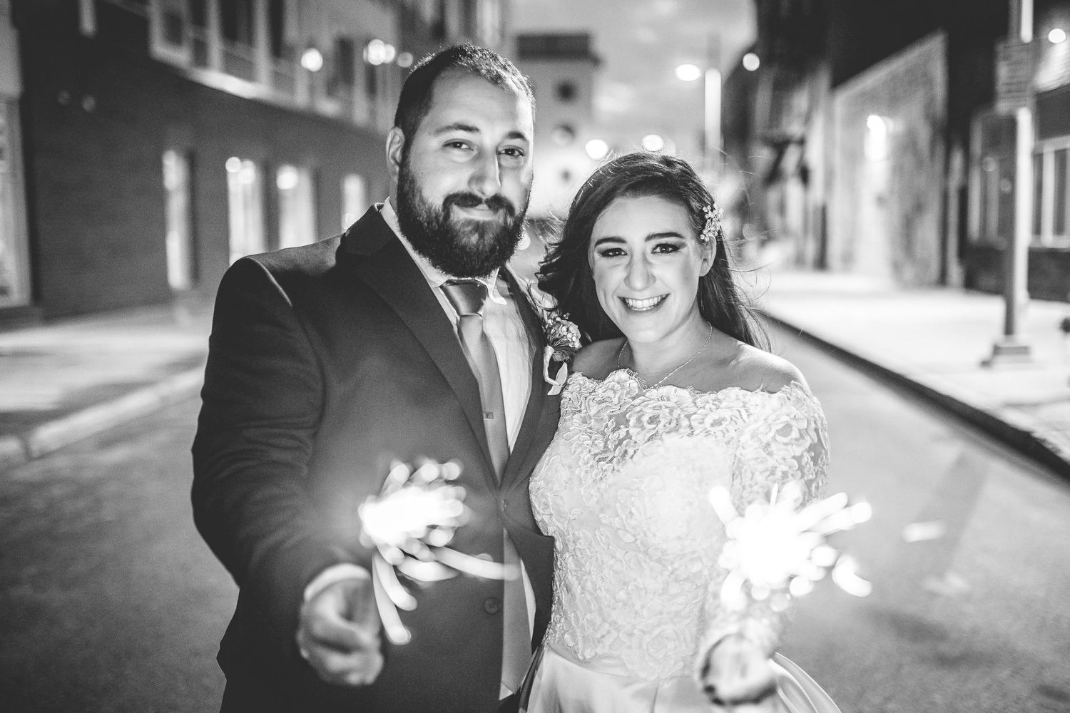 Couple poses with sparklers on dimly lit city street during wedding celebration.