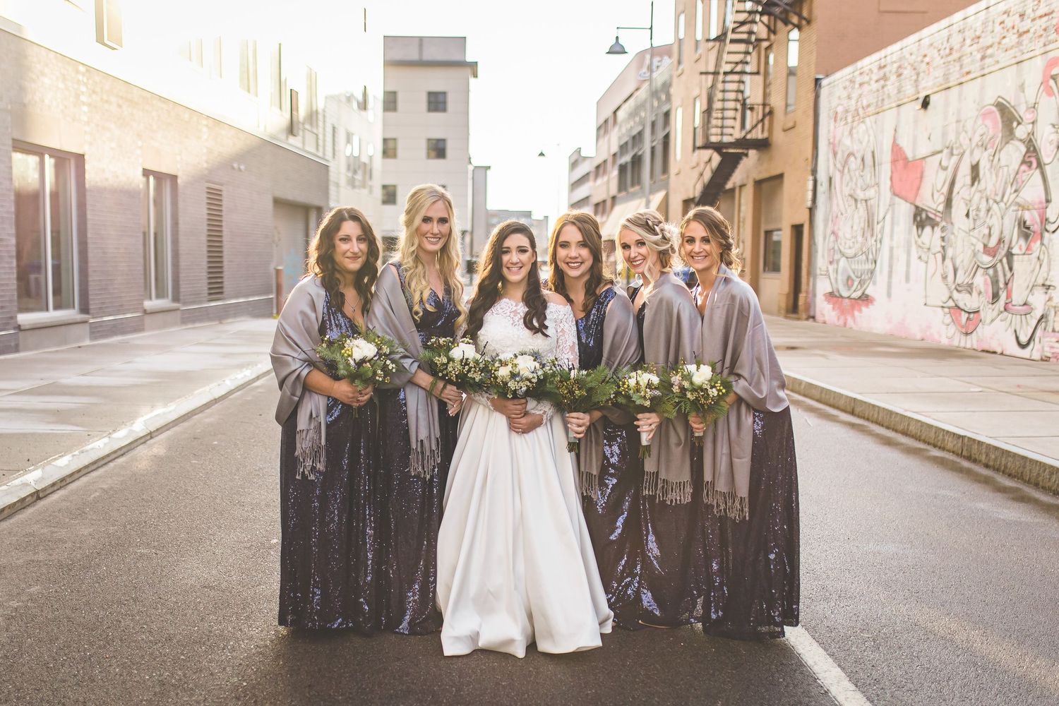 Wedding party in gray shawls poses together on urban street.