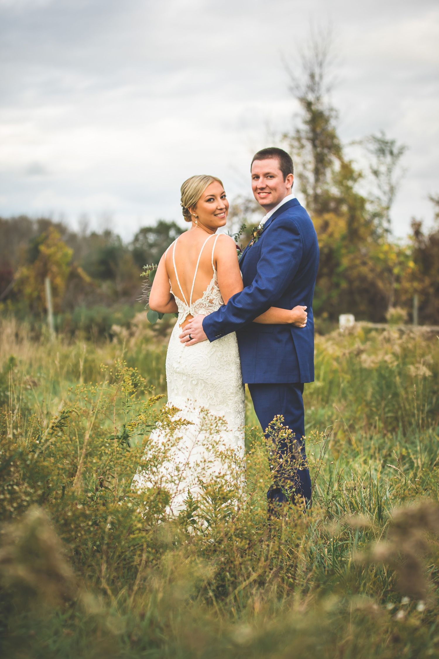 Newlyweds embrace while standing in tall grass during outdoor photo session.