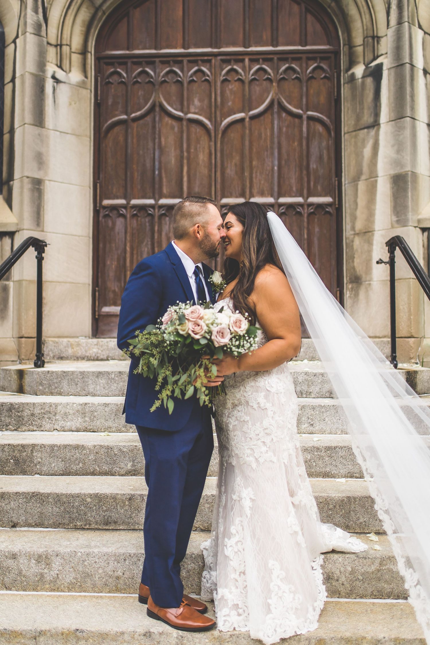 Romantic wedding portrait of couple embracing on stone church steps with ornate wooden door background.
