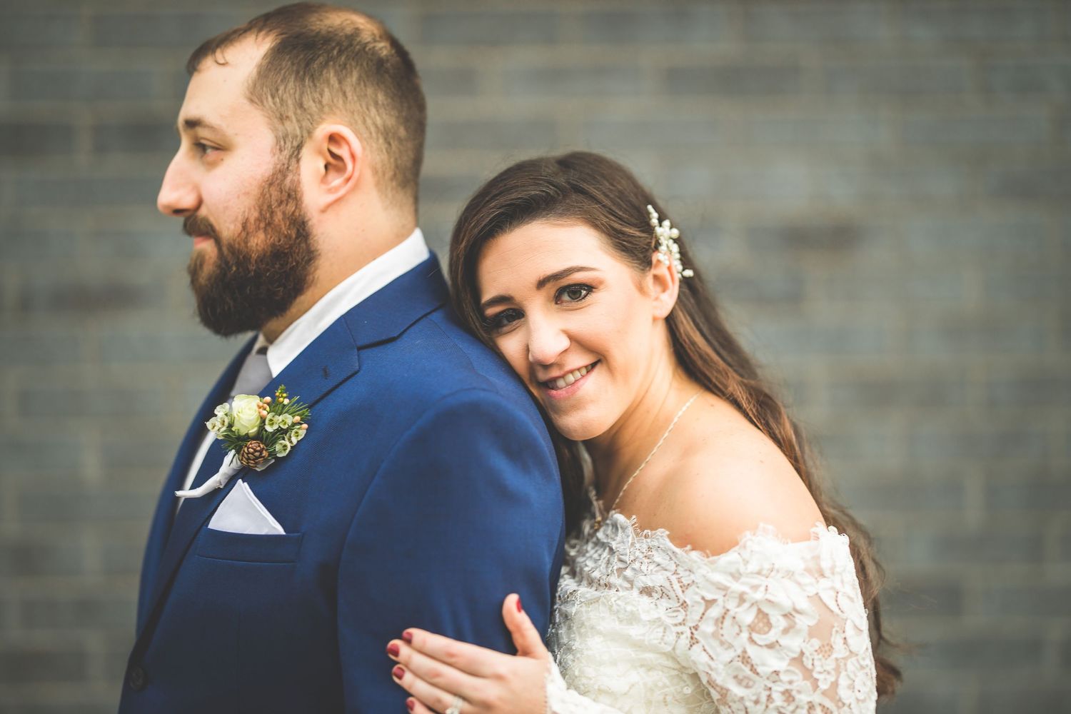 A romantic wedding portrait of a couple in a navy suit and lace dress against a brick wall.