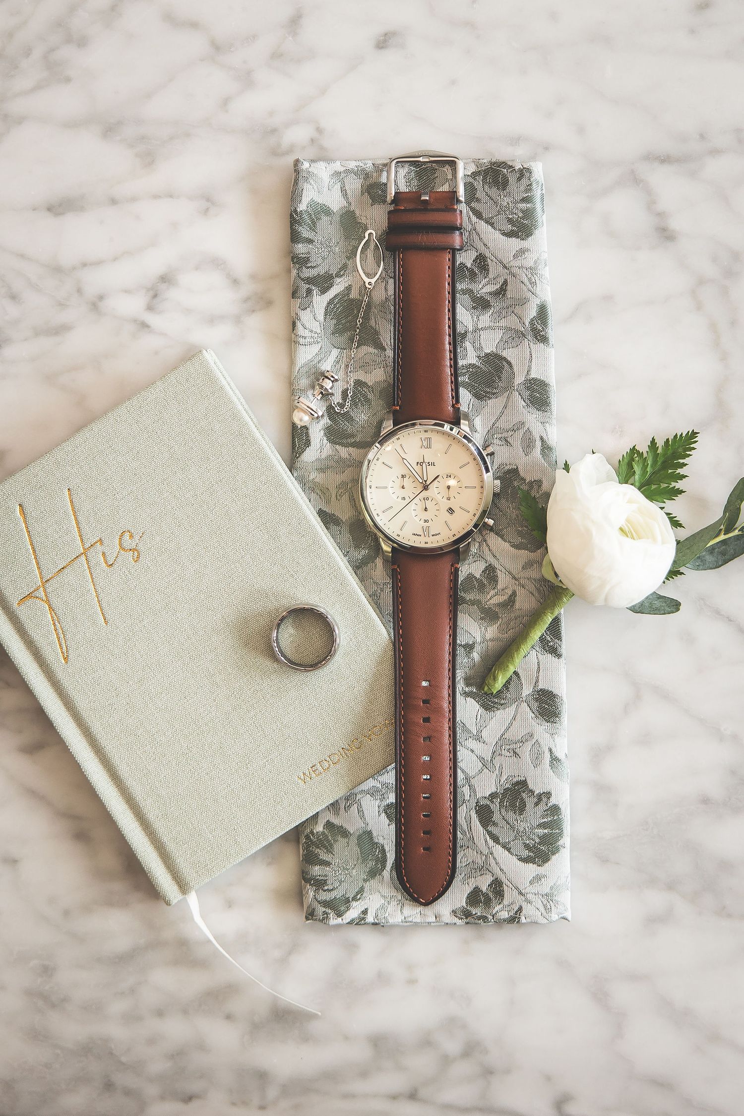 Wedding details including a leather watch, floral tie, and vow book arranged on marble surface.
