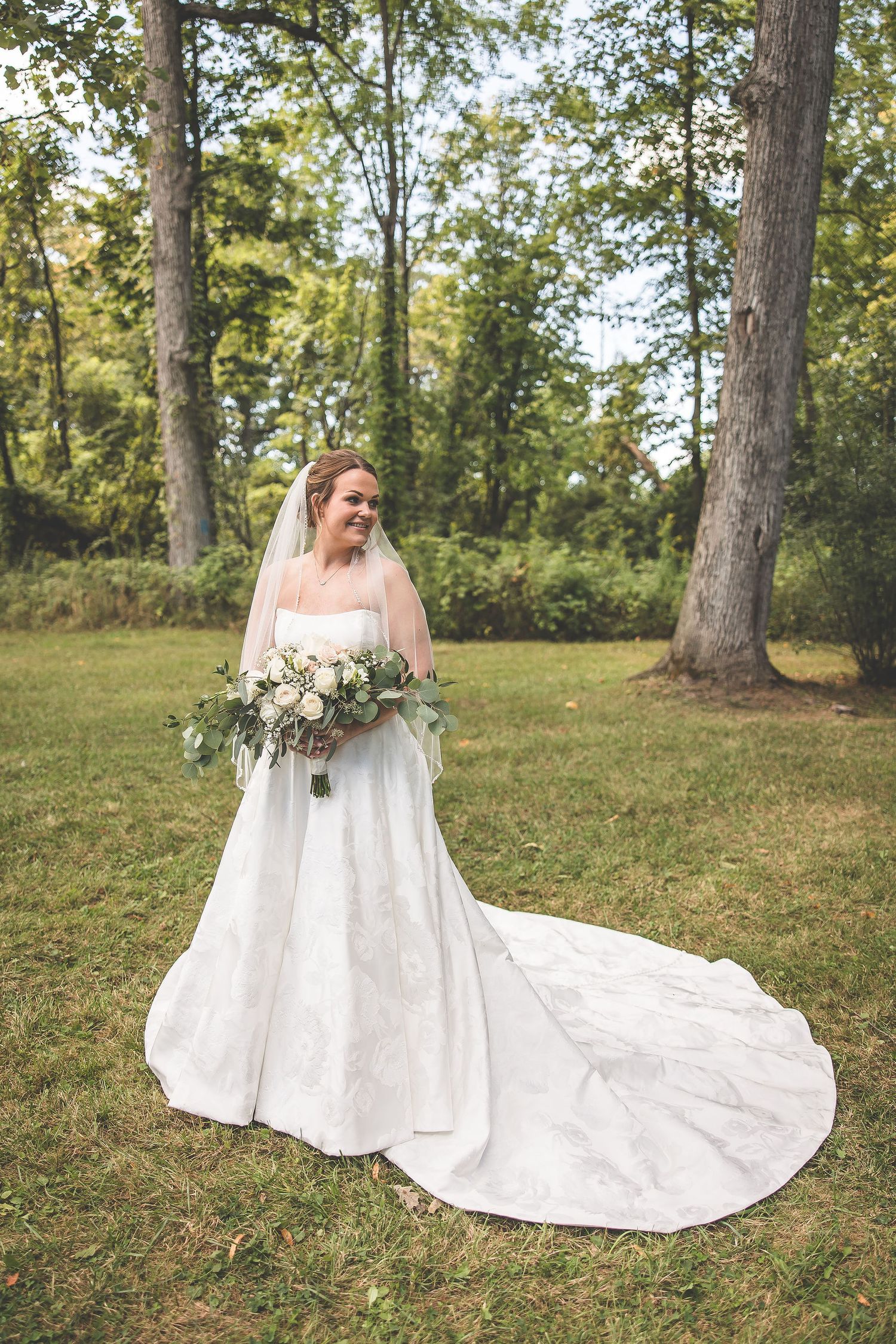 A white wedding dress with long train spreads across green grass in a wooded outdoor setting.