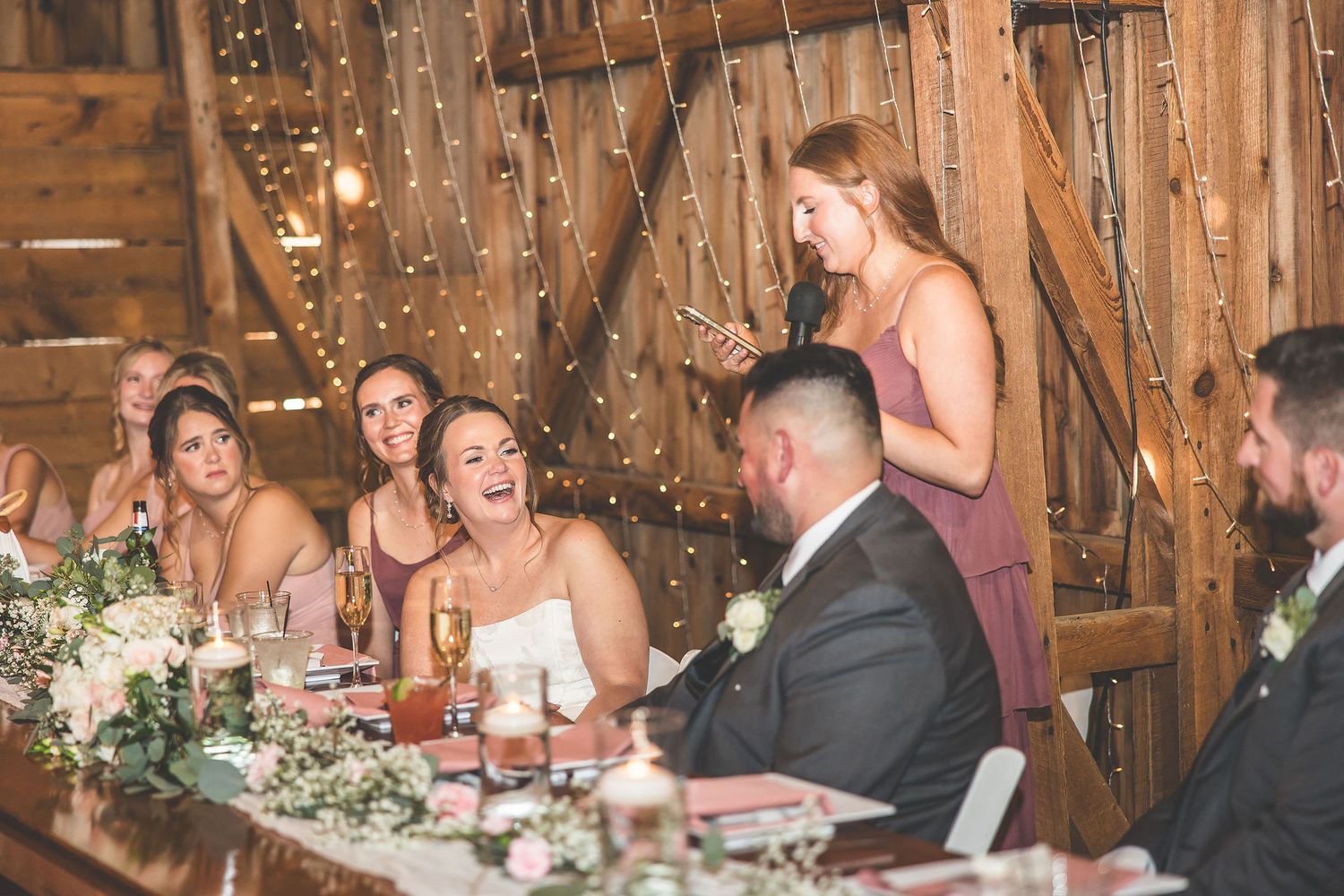 Wedding reception celebration with guests seated at decorated tables in rustic barn setting.