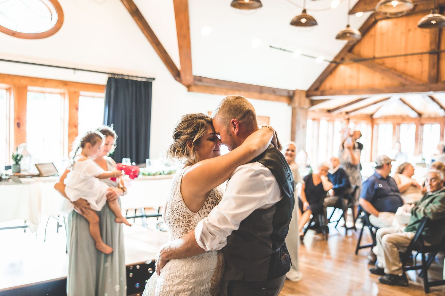Wedding guests dancing and celebrating at a rustic indoor reception venue with natural lighting.