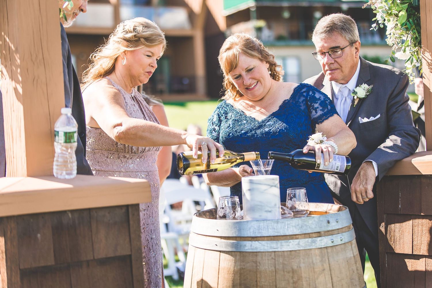 Wedding guests gather around a rustic wine barrel during an outdoor celebration.