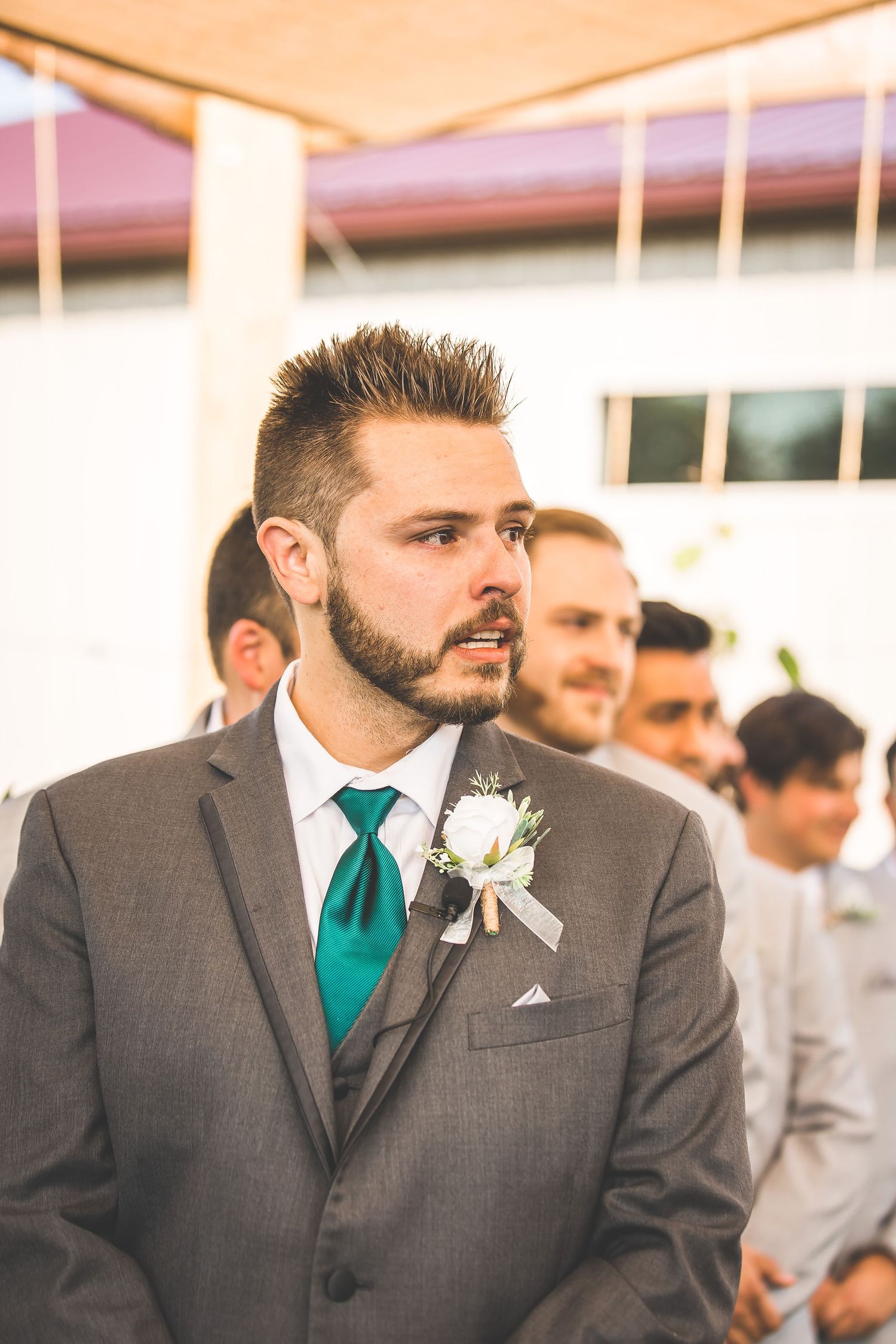 Groomsmen in charcoal gray suits with teal accents and white boutonnieres stand in a row at an outdoor wedding.