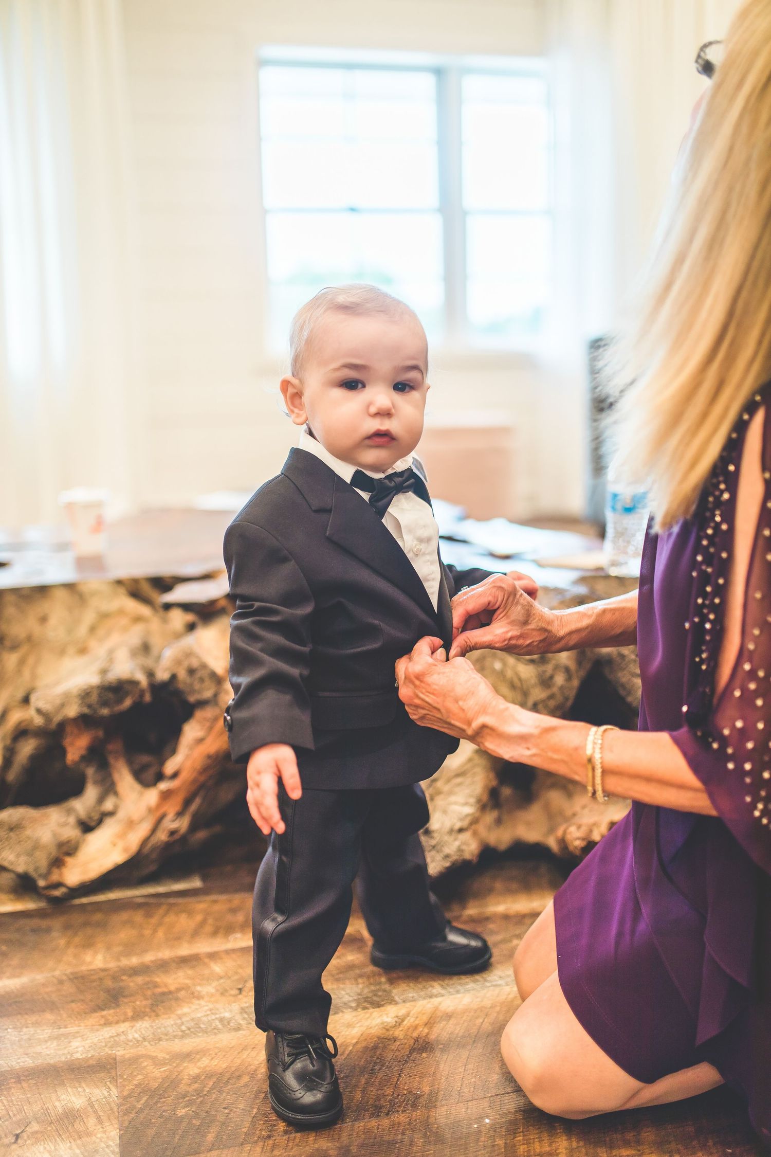 Small child in formal black suit stands on wooden floor during wedding preparations.