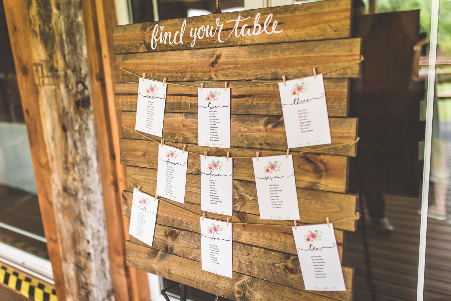 Wooden seating chart with cards hanging on strings against rustic wall.