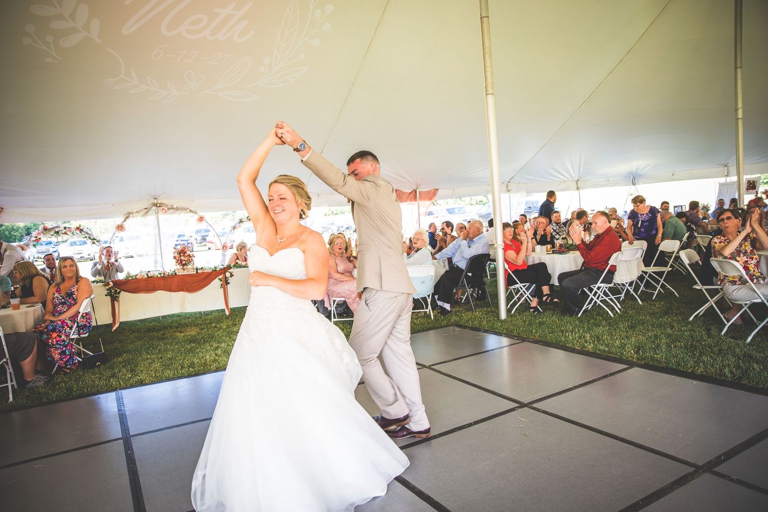Couple performs an energetic dance move during their wedding reception under a white tent.