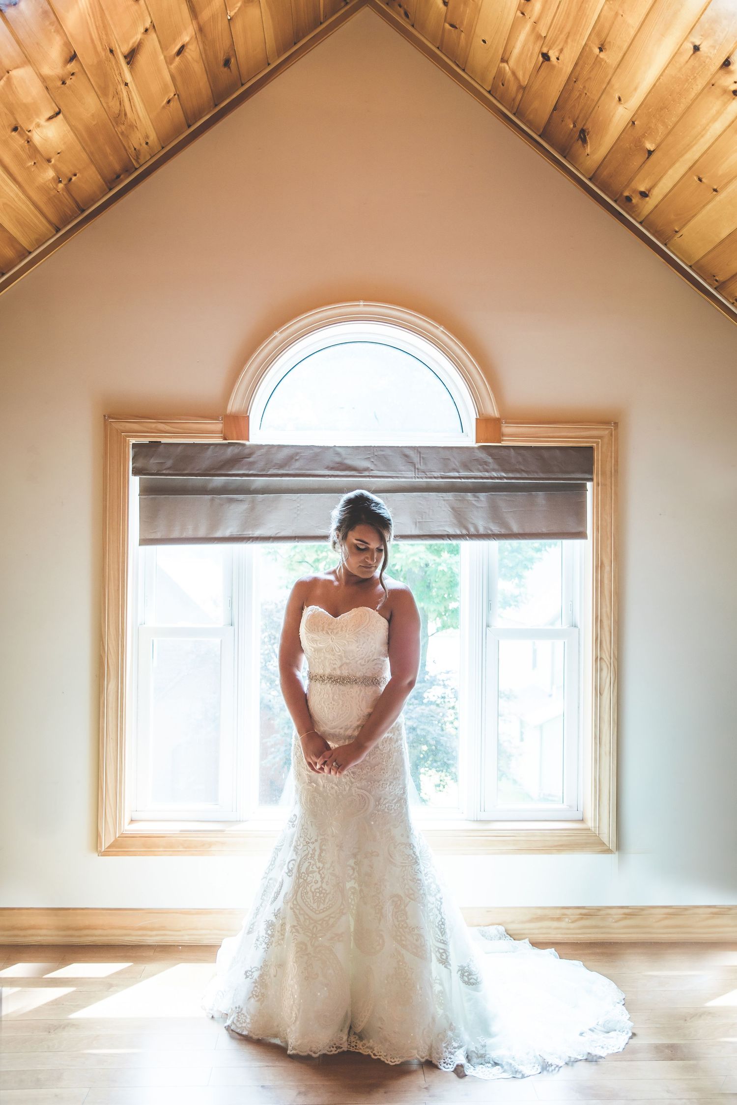 A white lace wedding dress is showcased in a rustic wooden room with natural window lighting.