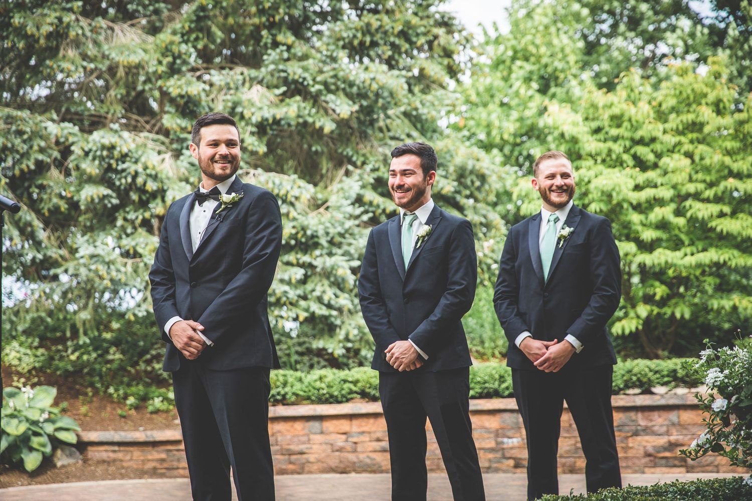 Three groomsmen in black tuxedos stand together outdoors during a wedding ceremony.