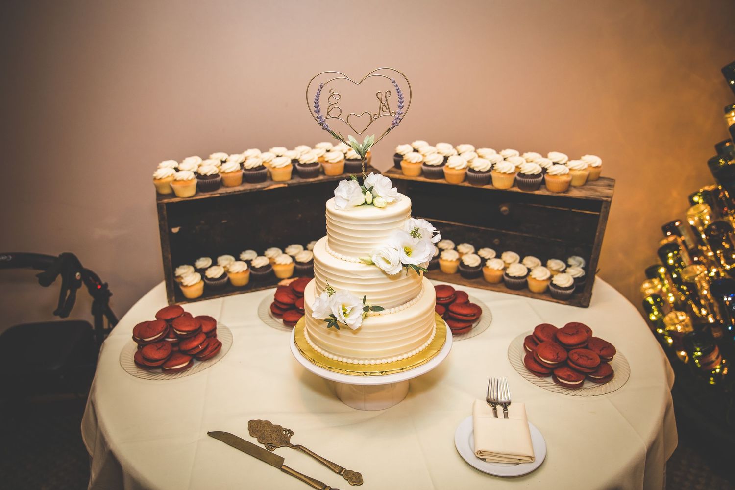 Wedding cake display with cupcakes and rustic decorations on white tablecloth.