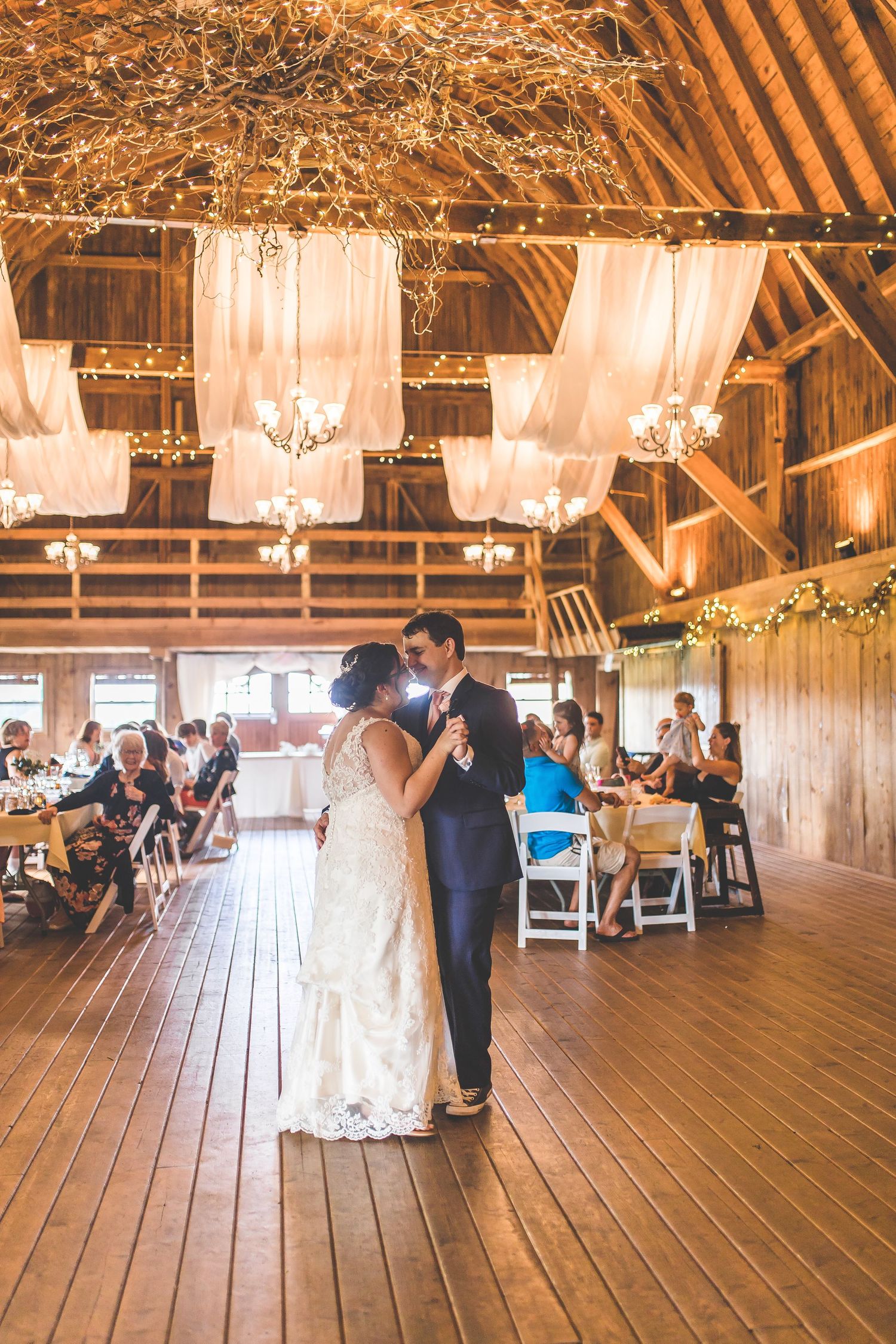 Newlyweds dance together on wooden floor of rustic barn venue with string lights and white decor.