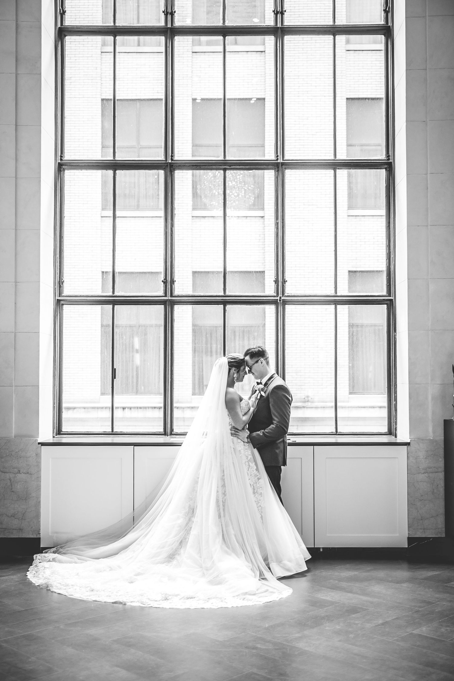 Black and white portrait of wedding couple standing by large geometric window.