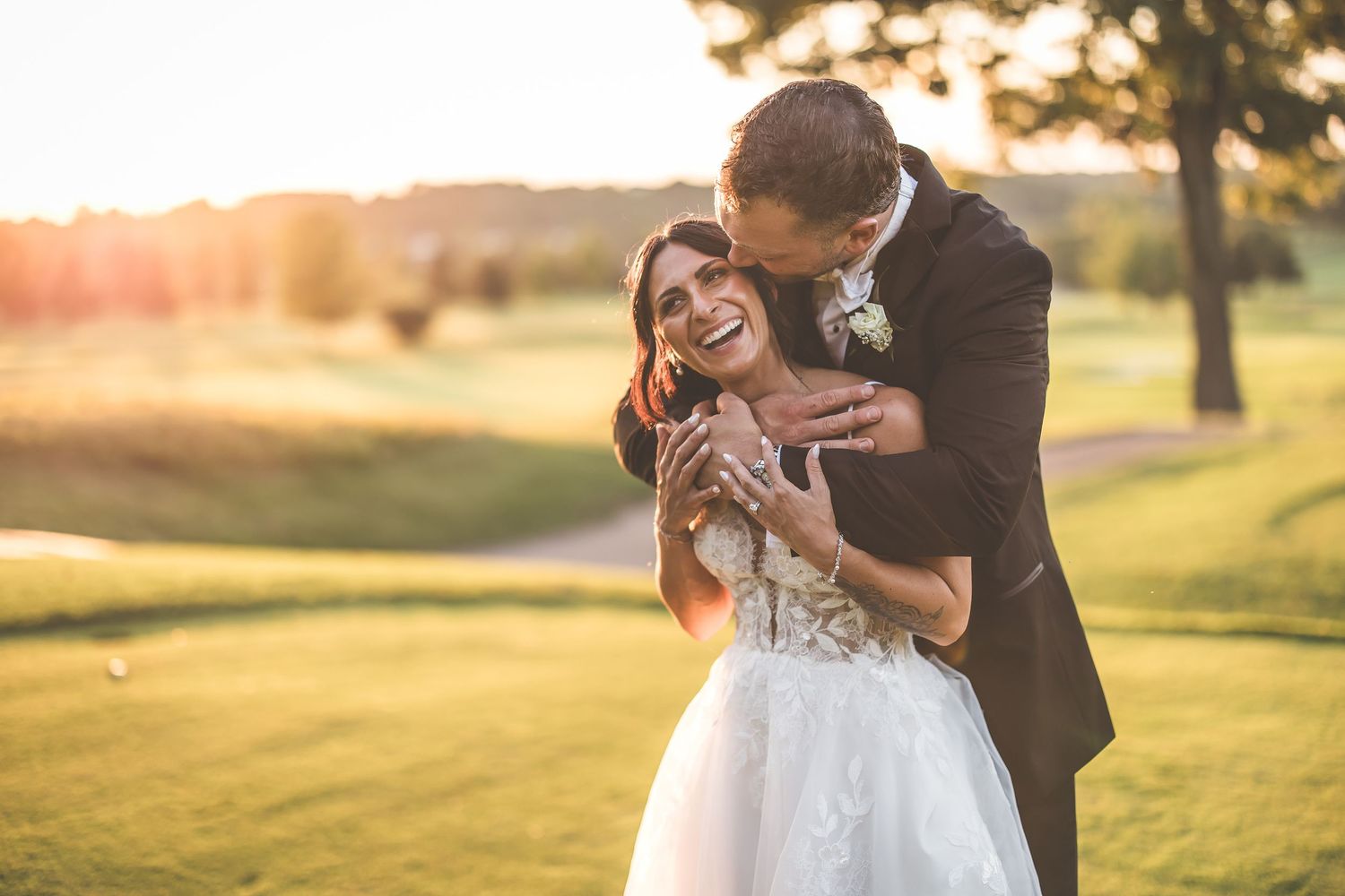Joyful bride in lace gown laughing as groom in black suit embraces her during golden hour outdoor wedding.
