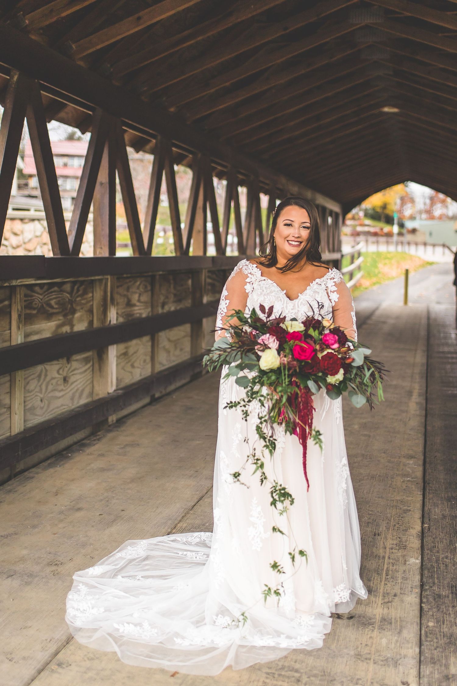 Bride in white lace gown poses with cascading bouquet inside rustic covered bridge.