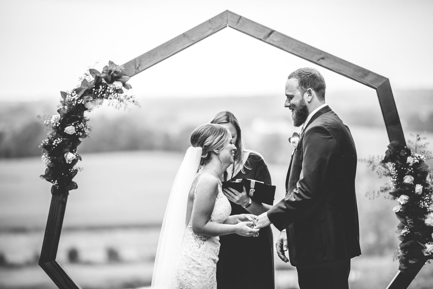 Black and white capture of wedding ceremony under geometric arch with natural landscape.
