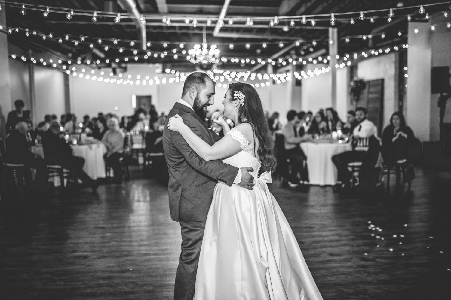 Couple dances beneath twinkling lights in an industrial-style wedding venue.