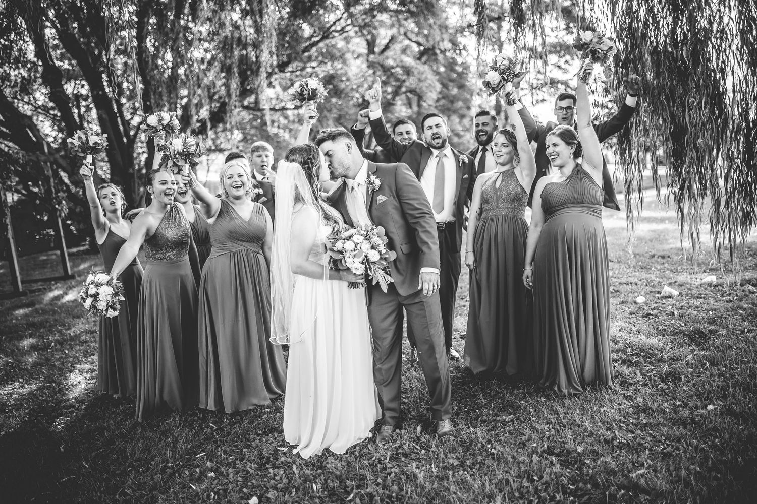 Black and white photo of wedding party gathered under trees for group portrait.