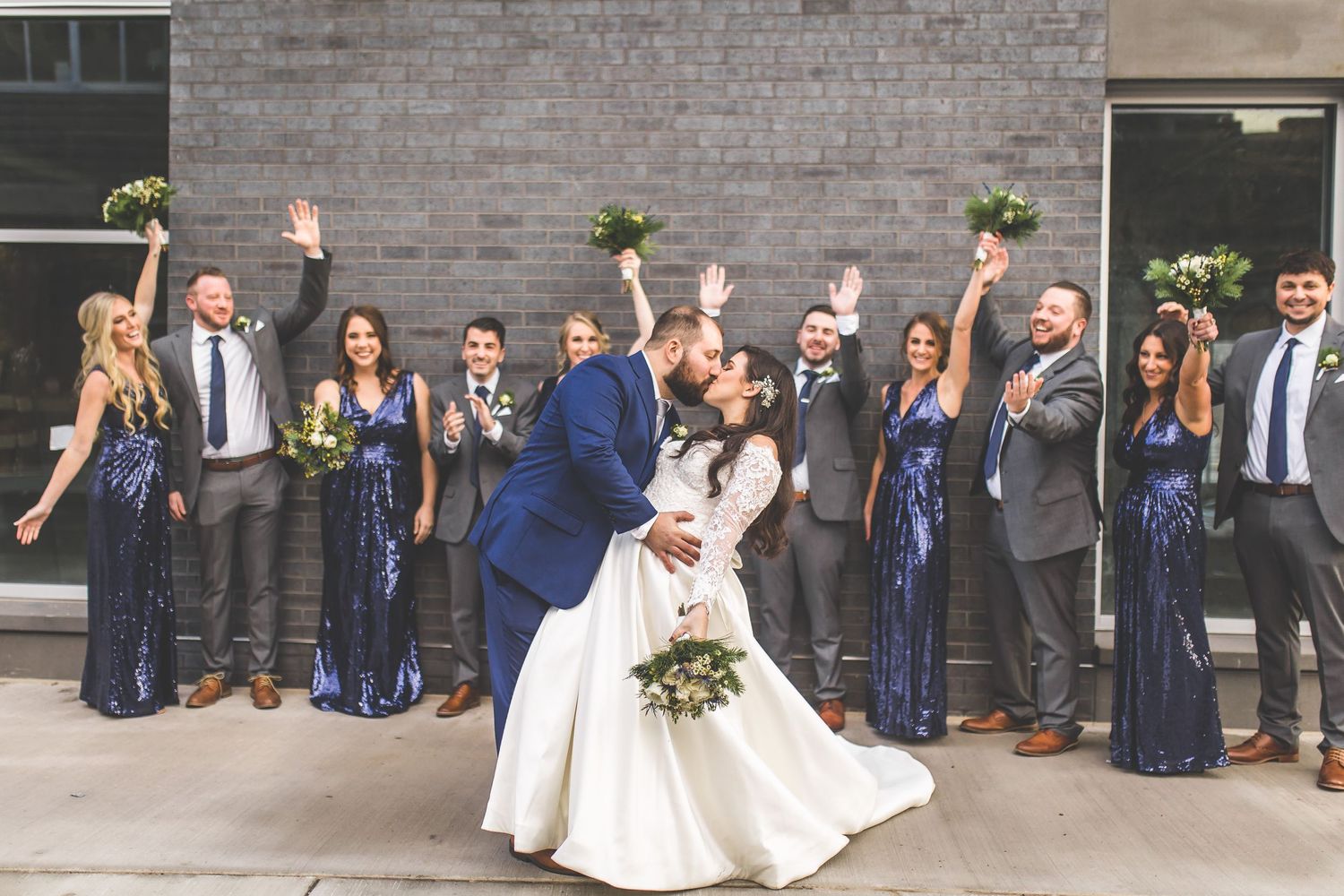 Wedding party celebrates with raised arms outside brick building.