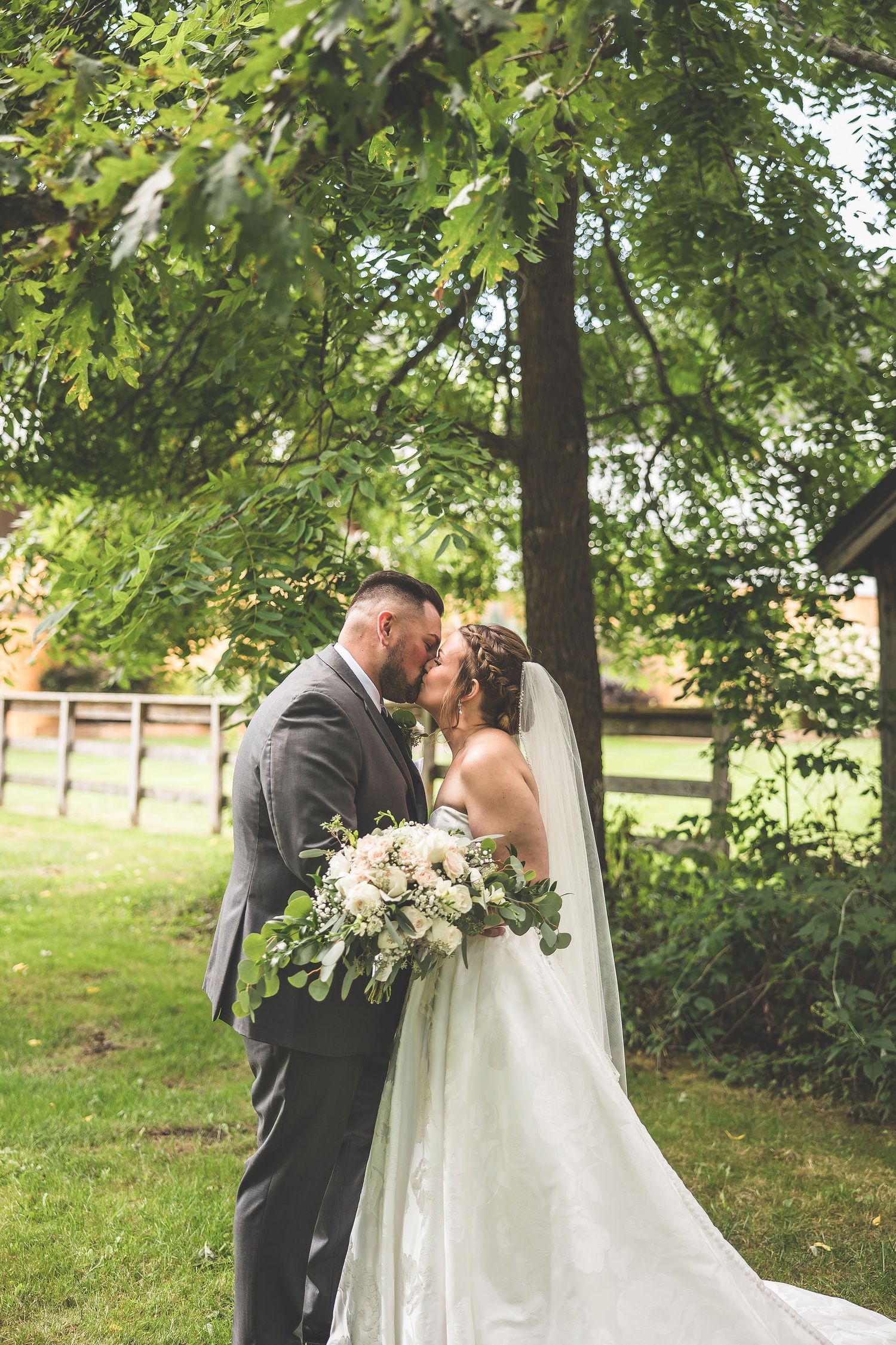 A romantic outdoor wedding moment with a couple sharing a kiss under green trees near a rustic barn.