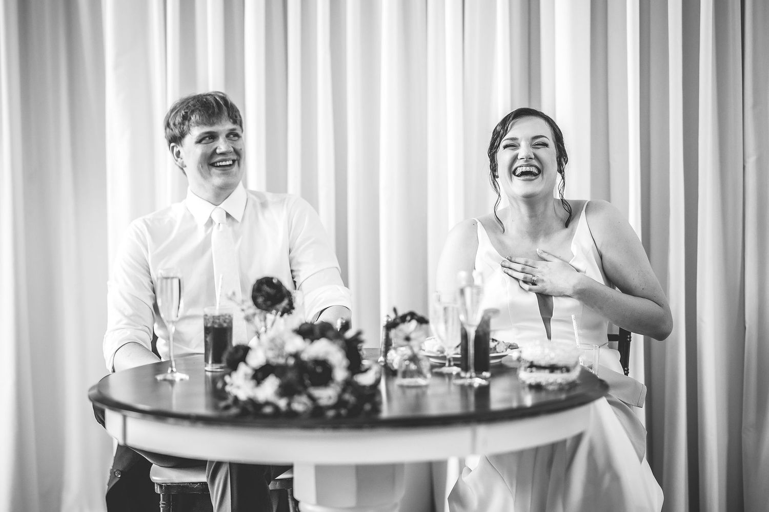 Wedding guests laugh and celebrate during a reception in an elegant venue with white drapes and round tables.