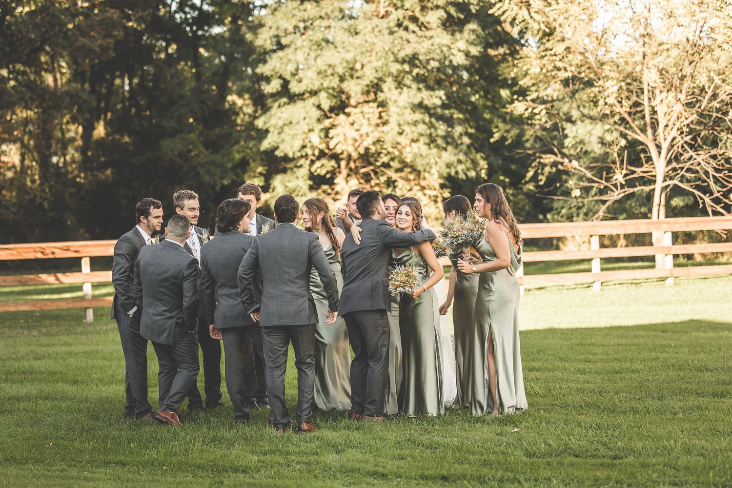 Wedding party members stand together in formal attire against a rustic fence and autumn backdrop.
