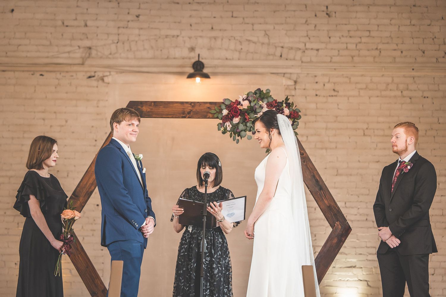 Wedding ceremony proceedings on wooden stage with exposed brick wall and white draping.