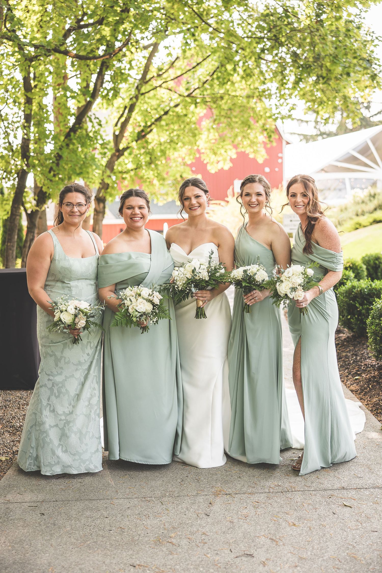 Bridesmaids in sage green dresses holding white bouquets pose together on garden path.