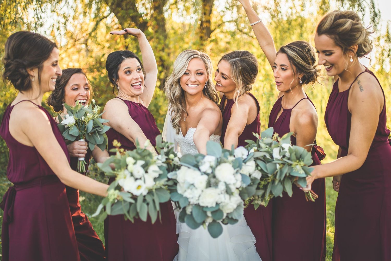 Bridesmaids in burgundy dresses celebrate with the bride holding eucalyptus bouquets during an outdoor autumn wedding.