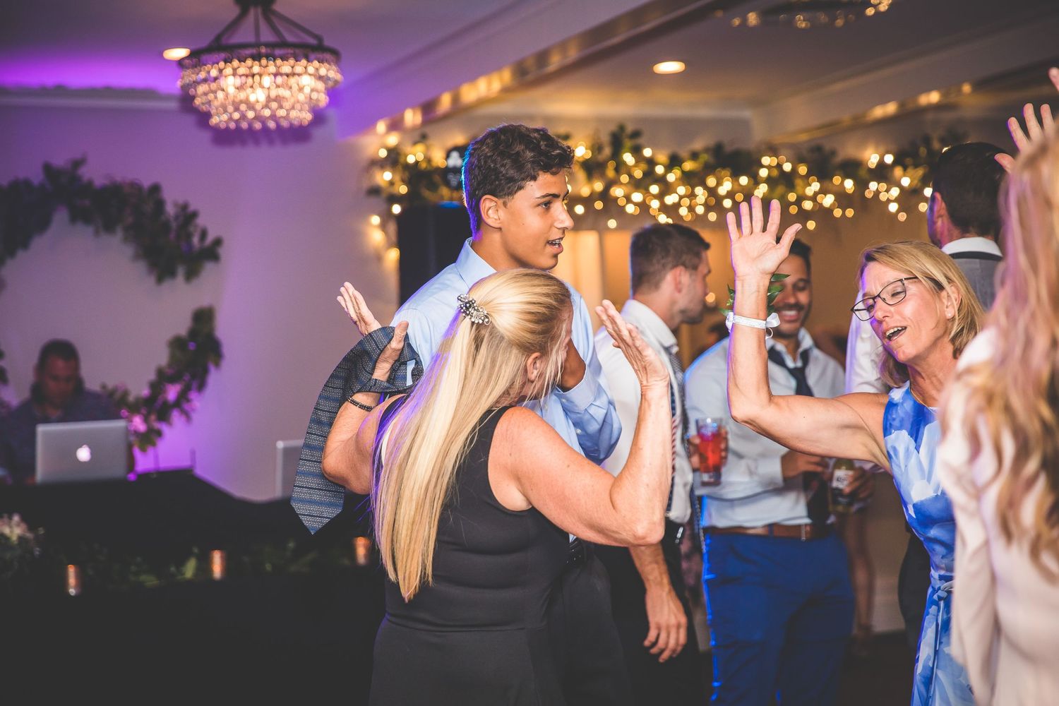 Wedding guests enjoying dance floor festivities under purple lighting and chandeliers.