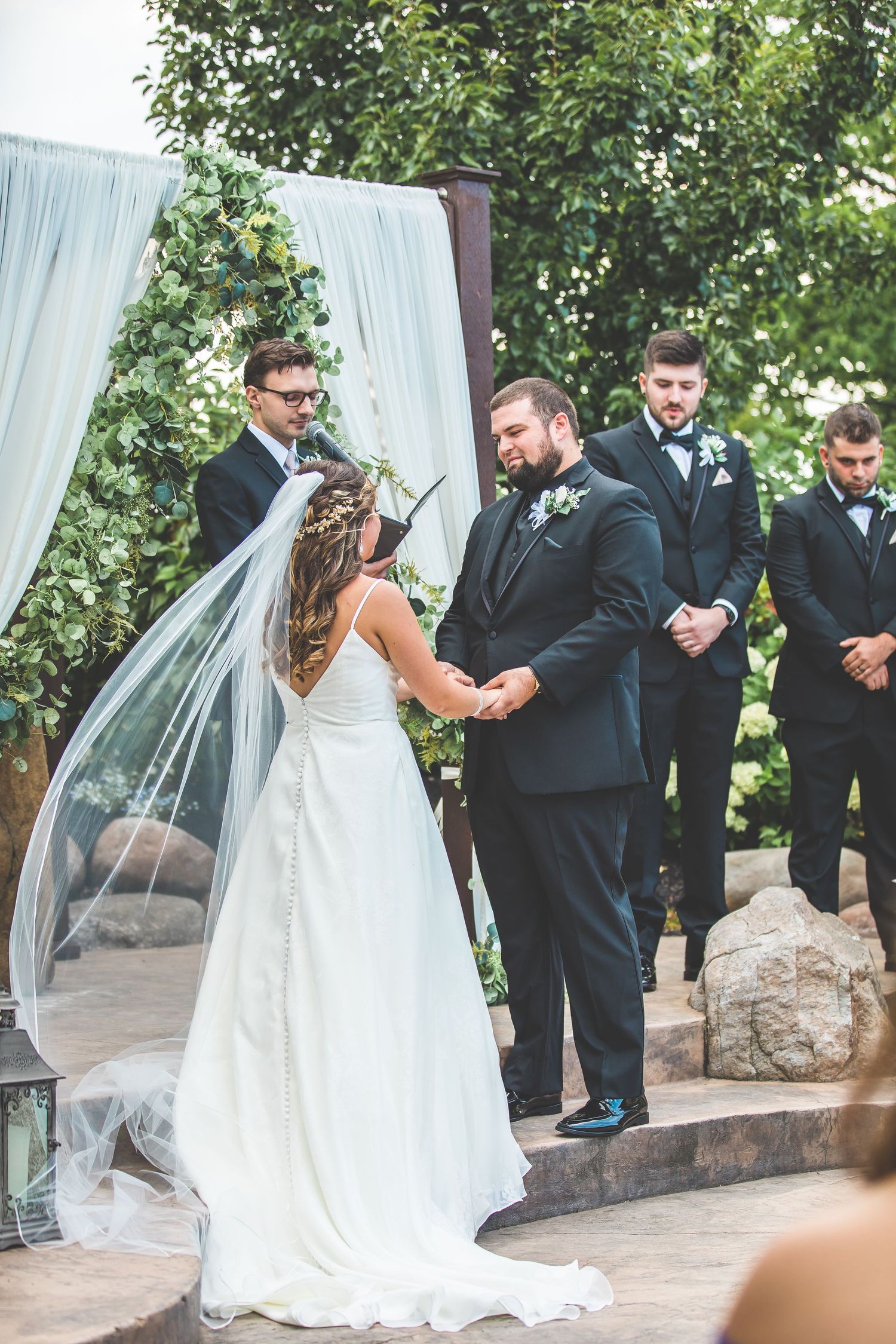 Wedding party in formal attire gathered outdoors near white draping.