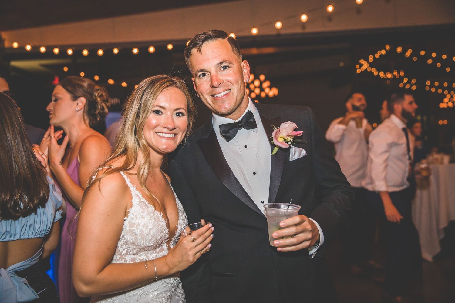 Wedding party celebrating on dance floor under warm string lights at reception venue.