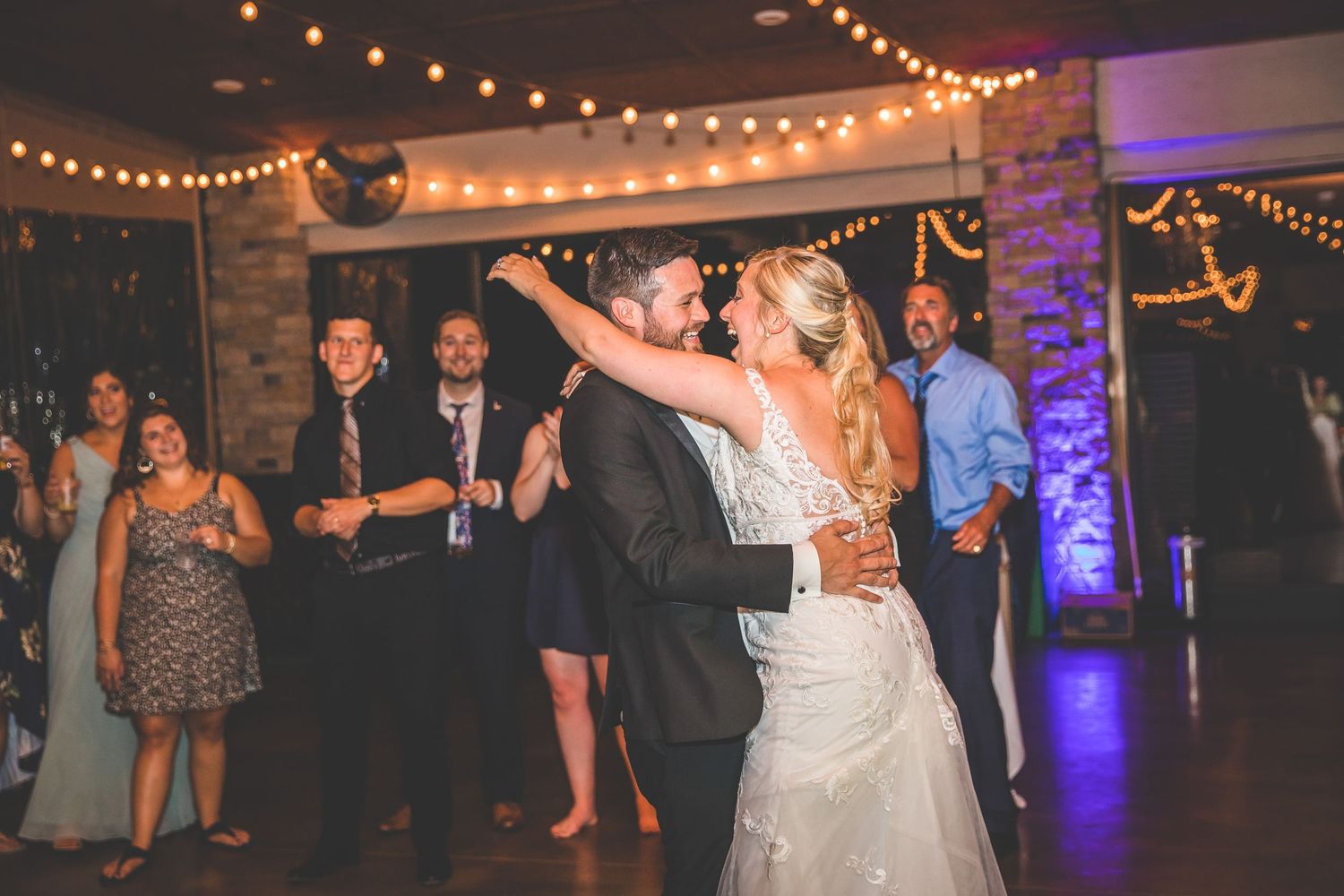 Newlyweds dance enthusiastically under string lights at their indoor reception.