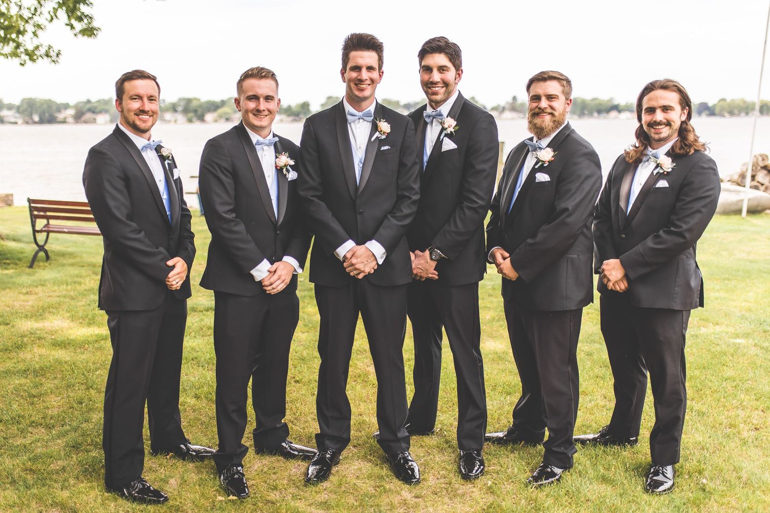 Groomsmen in black suits pose together on grass near waterfront.
