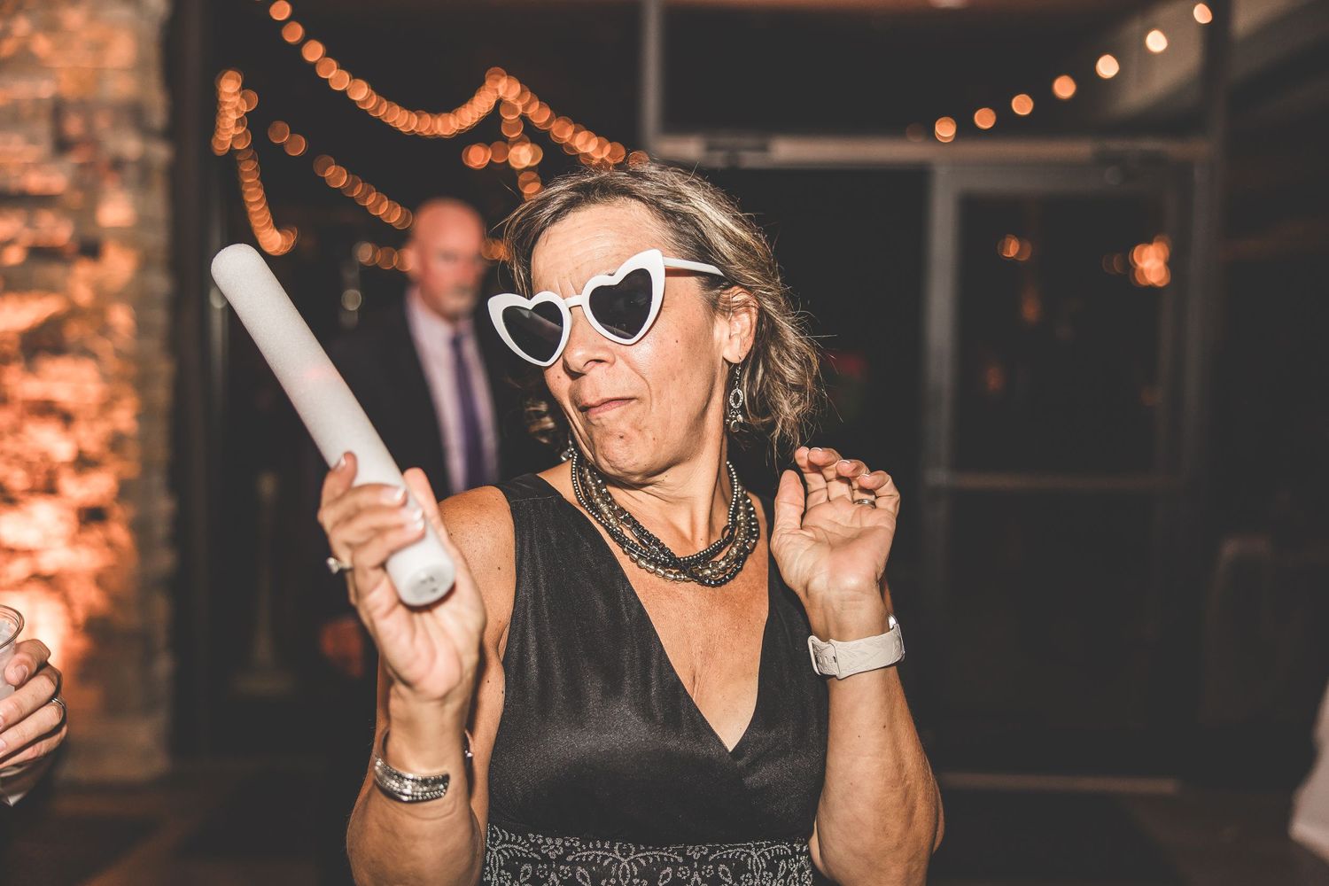 Wedding reception guests dancing and having fun under decorative string lights and brick walls.
