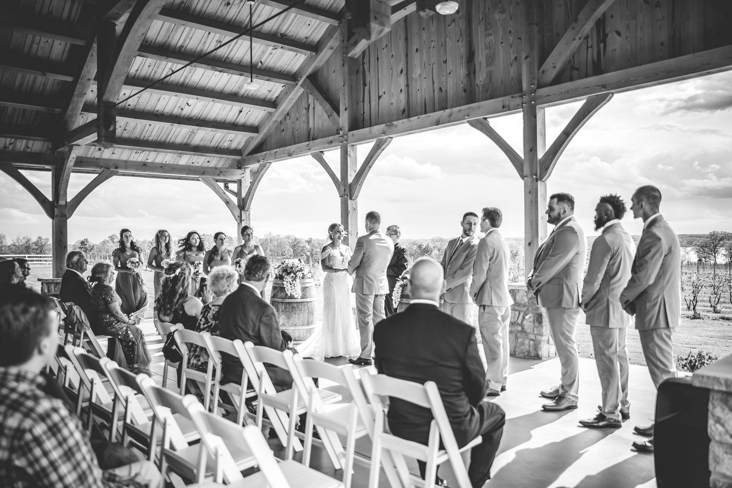 Black and white photo of wedding ceremony in rustic barn venue with exposed wooden beams.