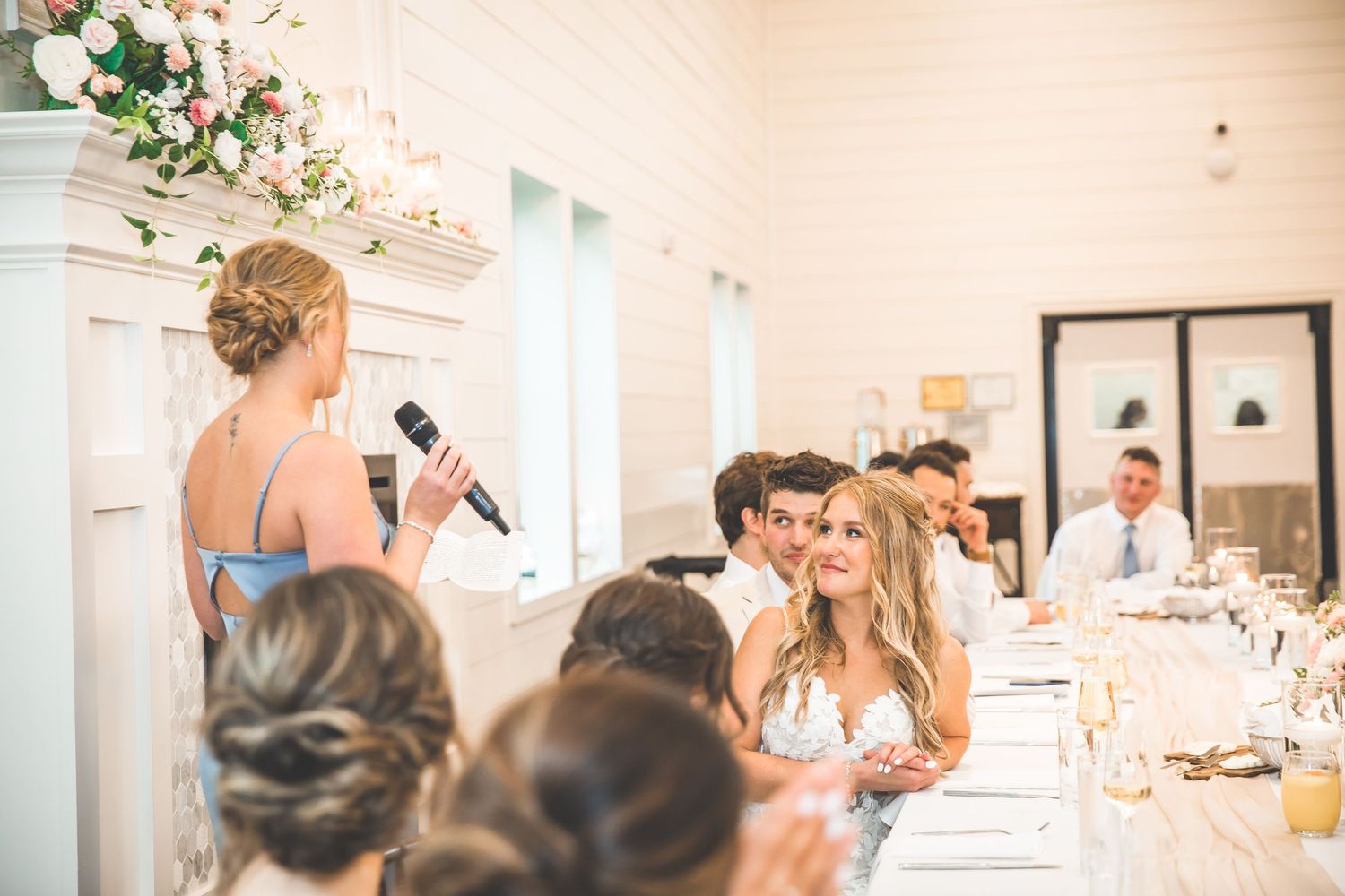 Wedding reception guests seated at elegant white tables with floral centerpieces in a bright indoor venue.